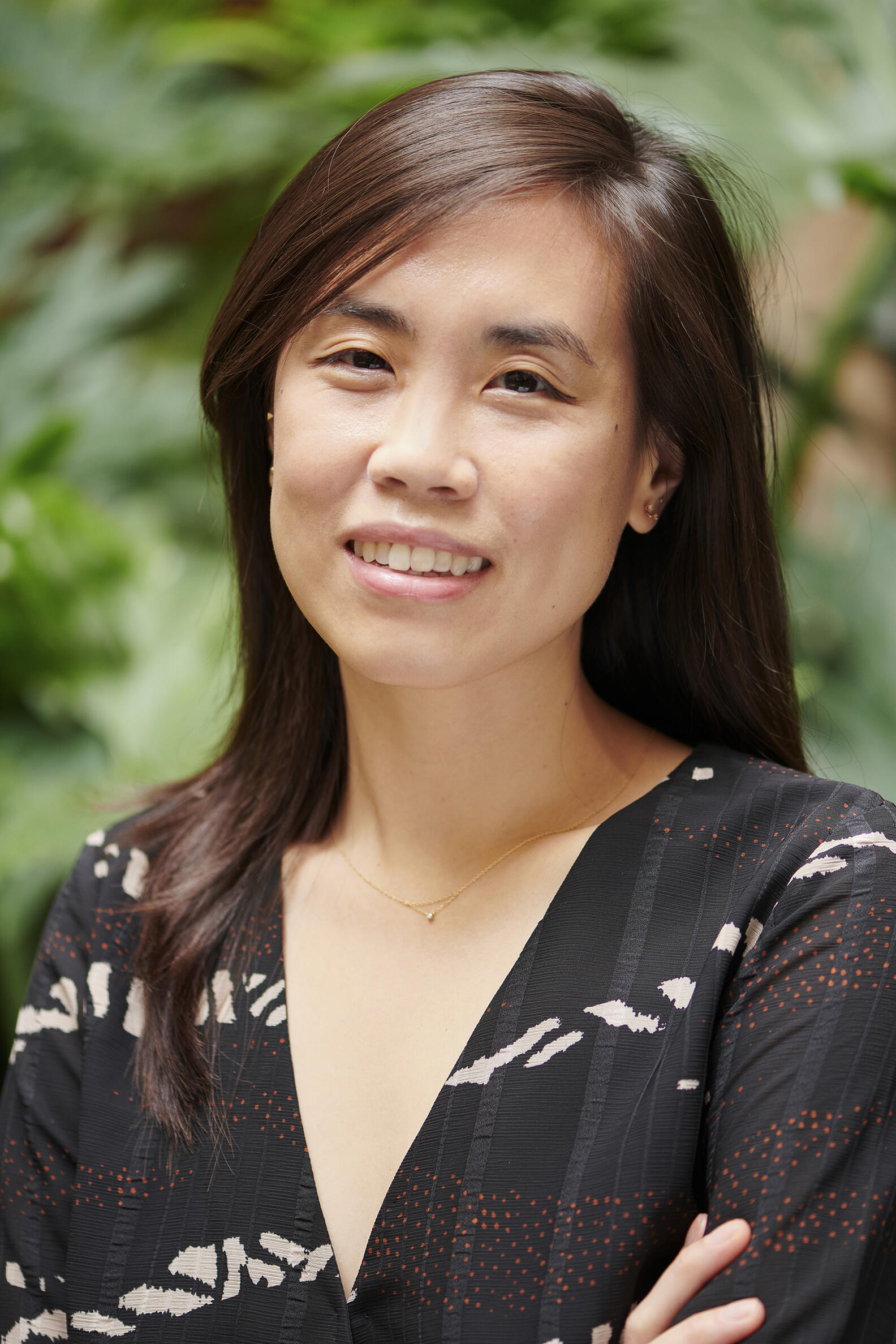 Young Asian woman with long, dark hair parted on the side, wearing a black patterned top with her arms crossed looking confidently at the camera. 