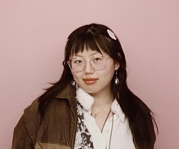Photo of a young Asian woman with long,dark hair, bangs, glasses, brown jacket and white shitrt looking at the camera with a slight smile 