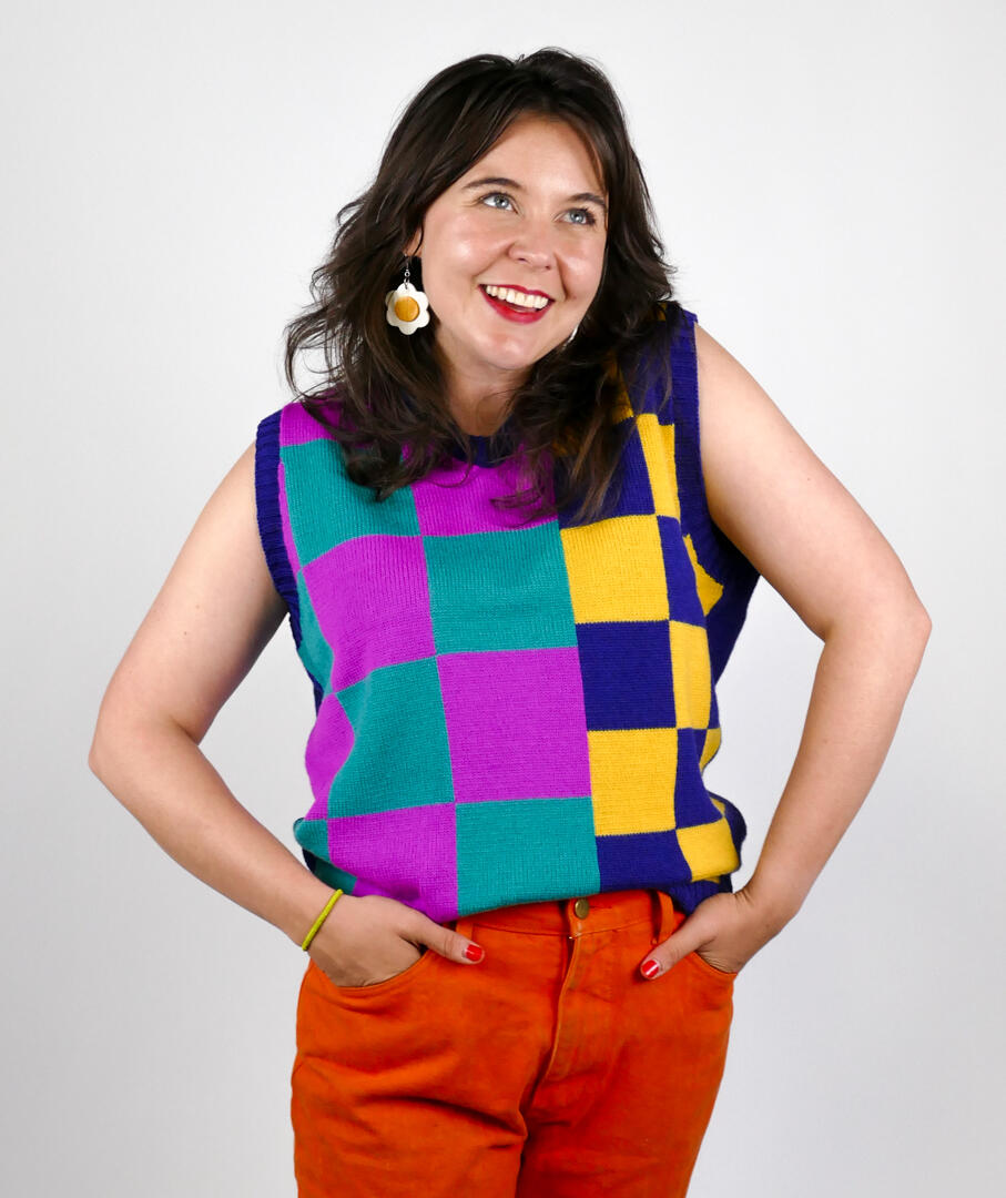 Young woman with shoulder length dark brown hair and bright red lipstick with a multicolored top with squares and orange slacks. 