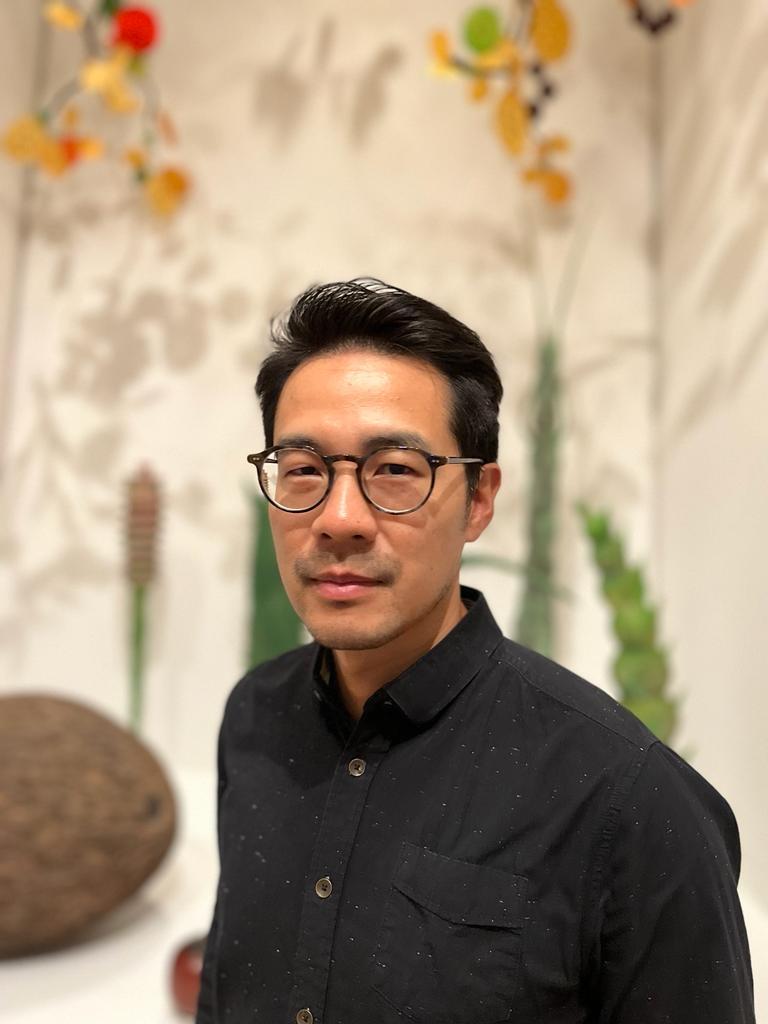 Asian man with short,dark hair, with glasses wearing a black button down shirt standing infront of oversized plants. 