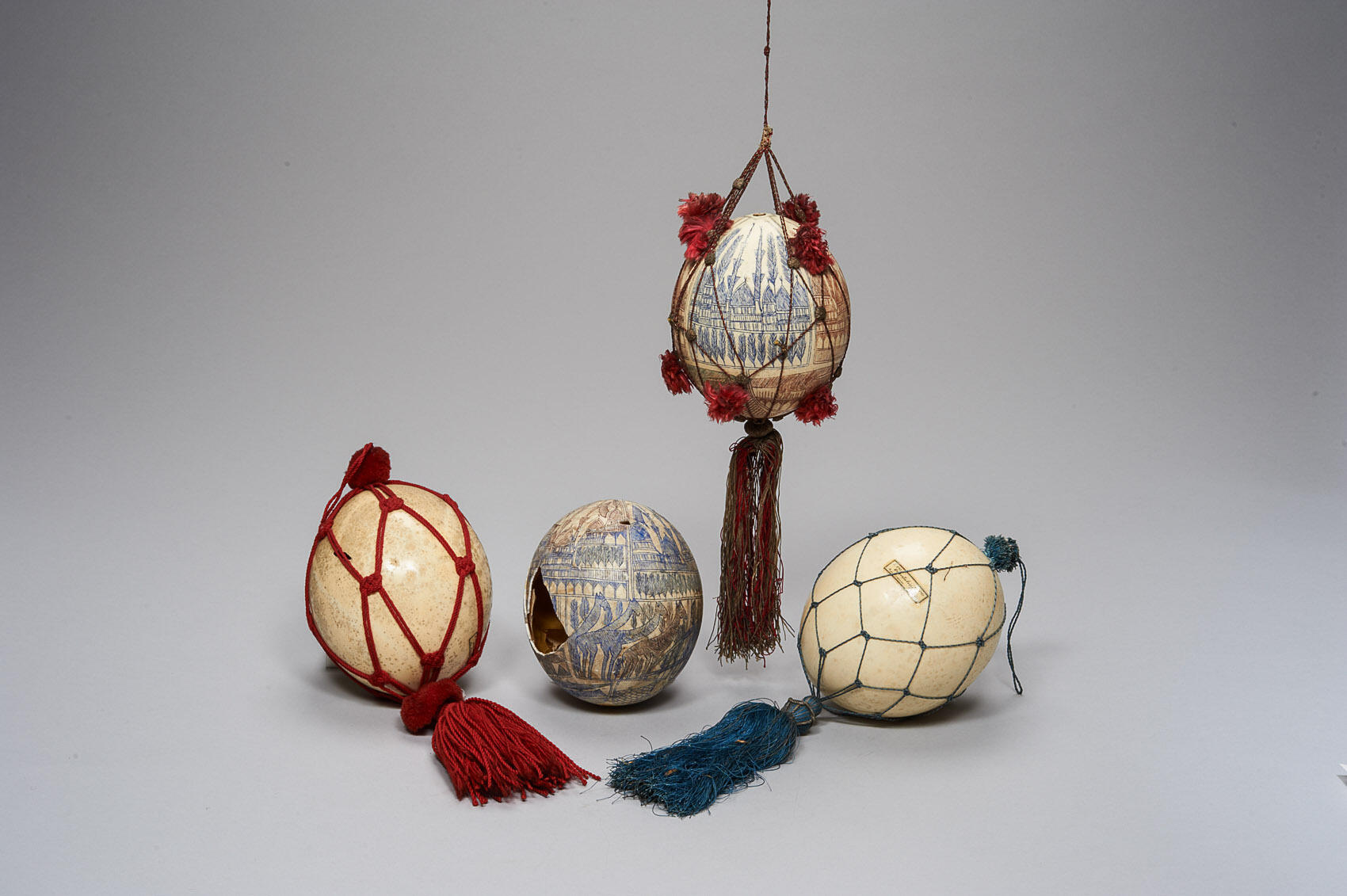 Four ostrich eggs, two with blue painted designs and two unpainted. Three of the eggs are encased by netting decorated with tassels and pom-poms.