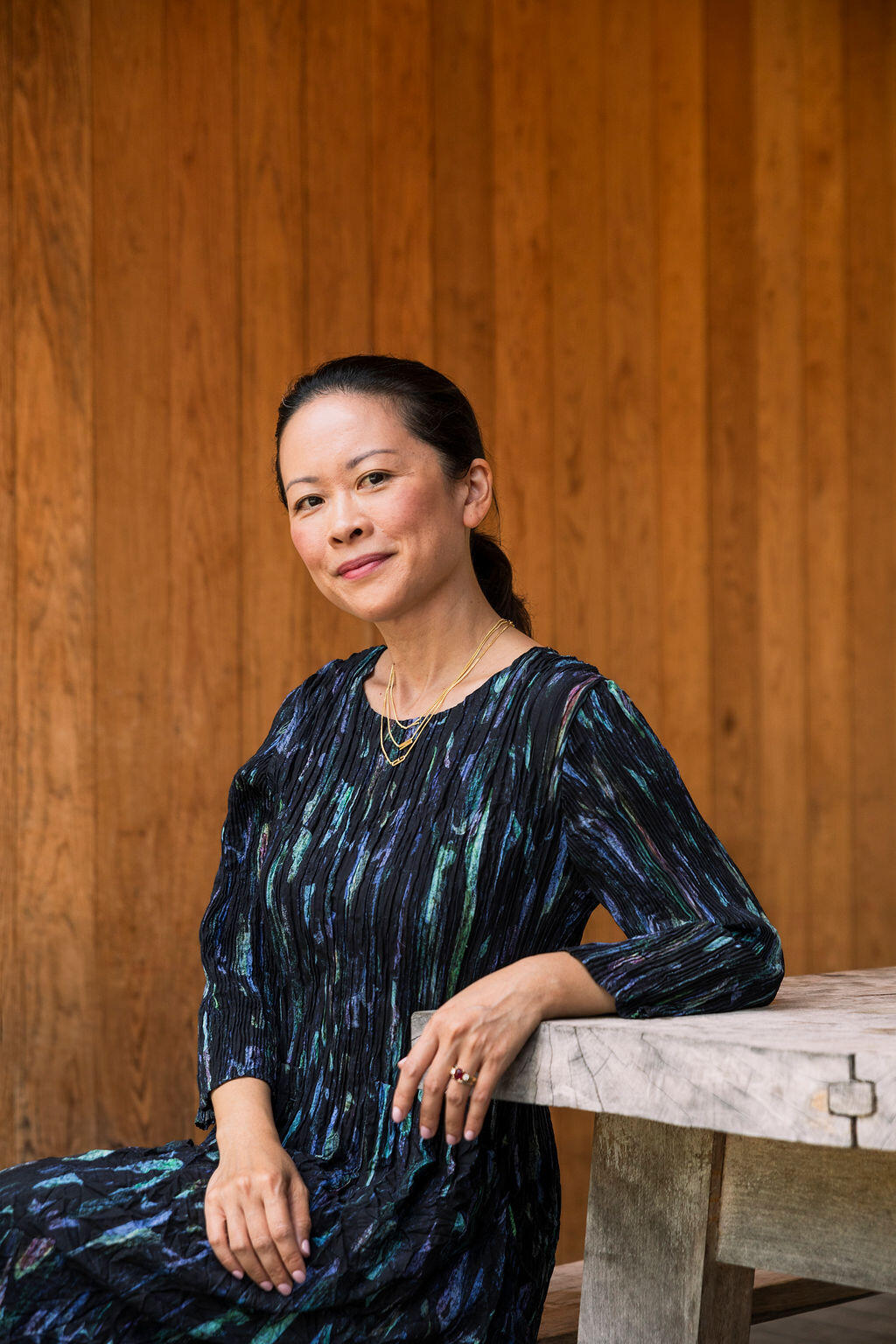 Seated Asian woman with long dark hair in a ponytail in a blue, patterned dress looking straight at the camera. 