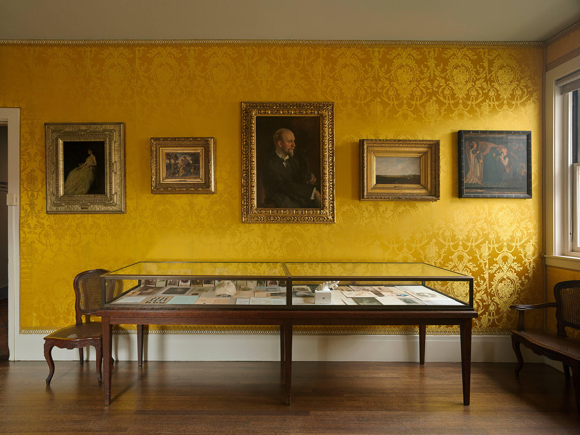 A half-length portrait of a white man holding a violin hanging on a yellow-upholstered wall with other paintings.