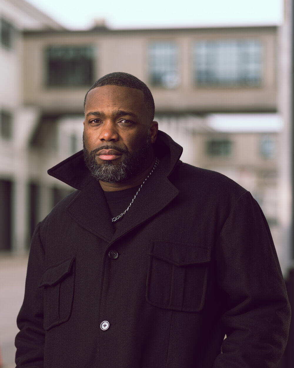 African American man with a beard and mustache wearing a black coat with the collar popped, looking directly at the camera. 
