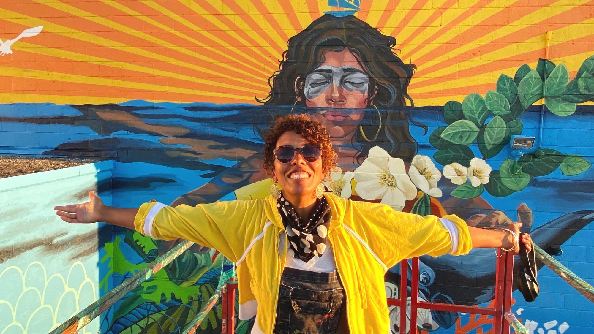 A hispanic woman in a bright yellow jacket and sun glasses, arms outstretched with sun glasses standing in front of building mural with a similar image behind her.  
