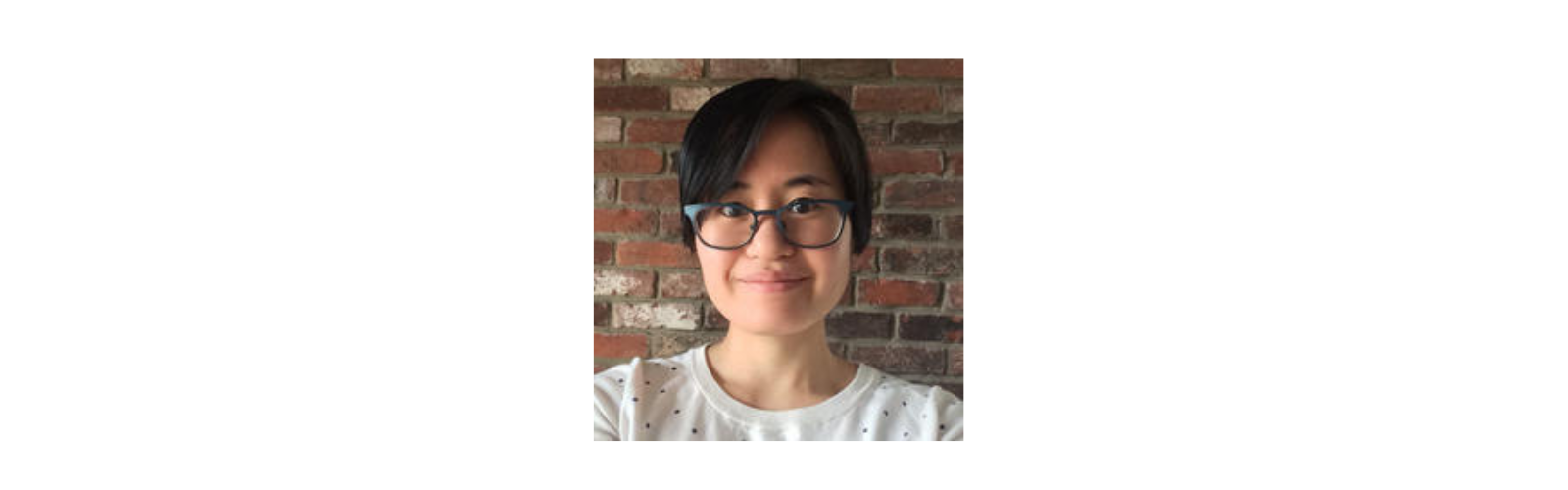 Young Asian woman whith short, dark hair with glasses standing against a brick wall. 