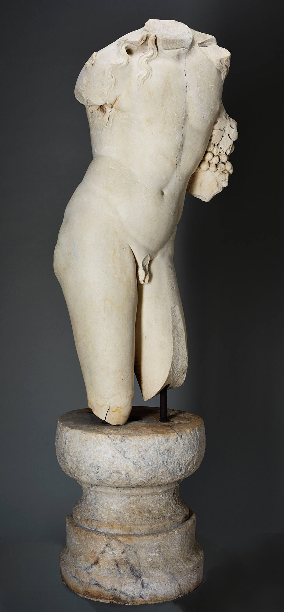  An off-white marble sculpture of a male torso including the upper part of the legs, with no arms or head, turned three-quarters to the right and holding grapes against the side of the chest.