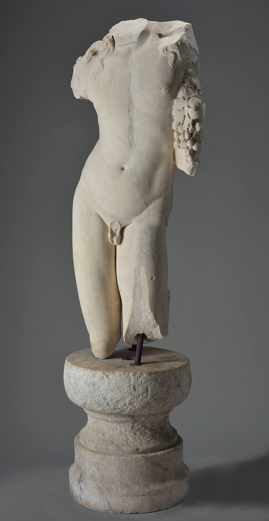 An off-white marble sculpture of a male torso including the upper part of the legs, with no arms or head, facing forwards and holding grapes against the side of the chest.