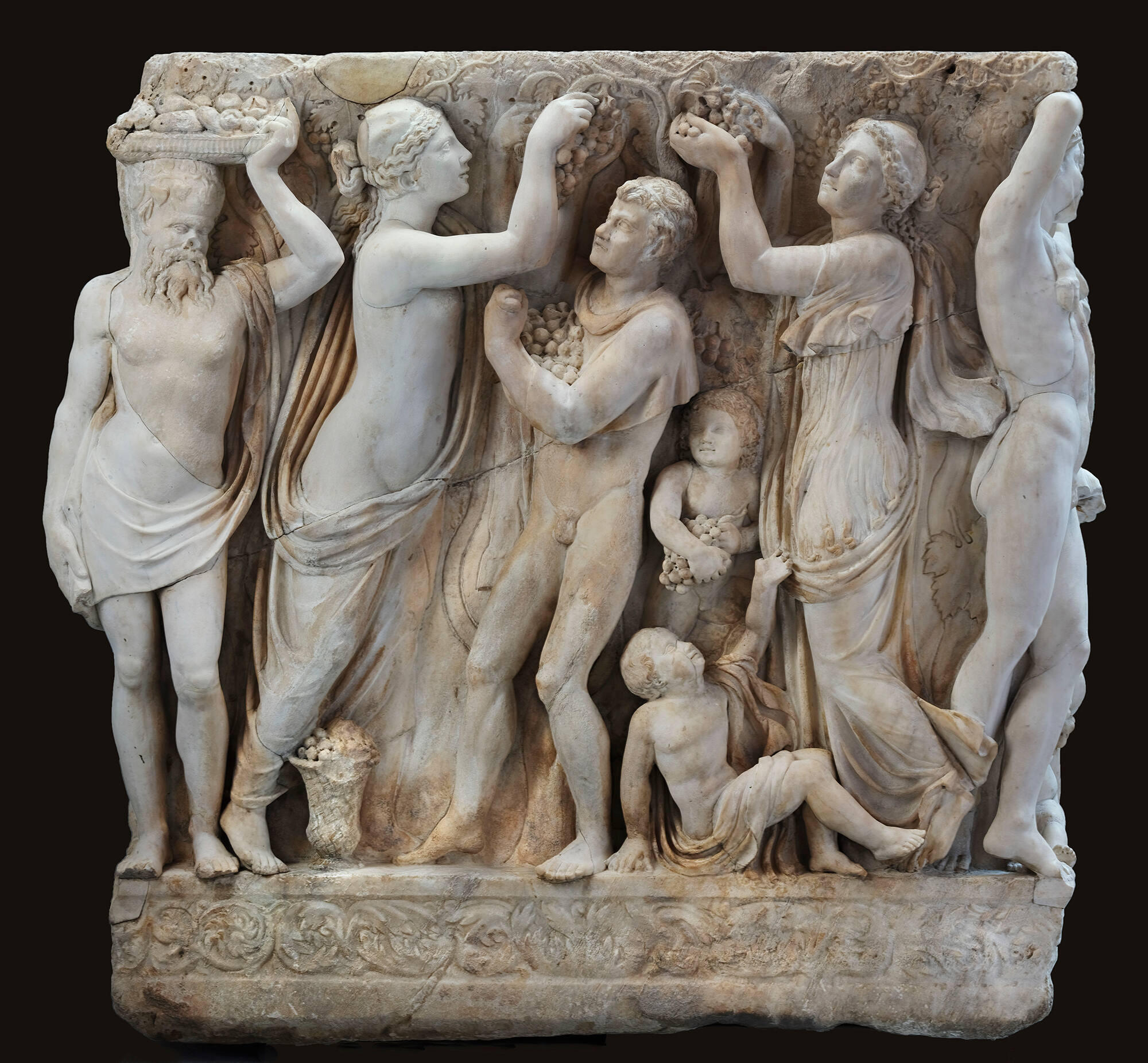  A high relief marble sculpture framed on the square end panel of a sarcophagus depicting maenads and satyrs interacting in various states of undress underneath grape vines, cupids, and a bald and bearded satyr on the left corner holding a basket of fruit on his head.