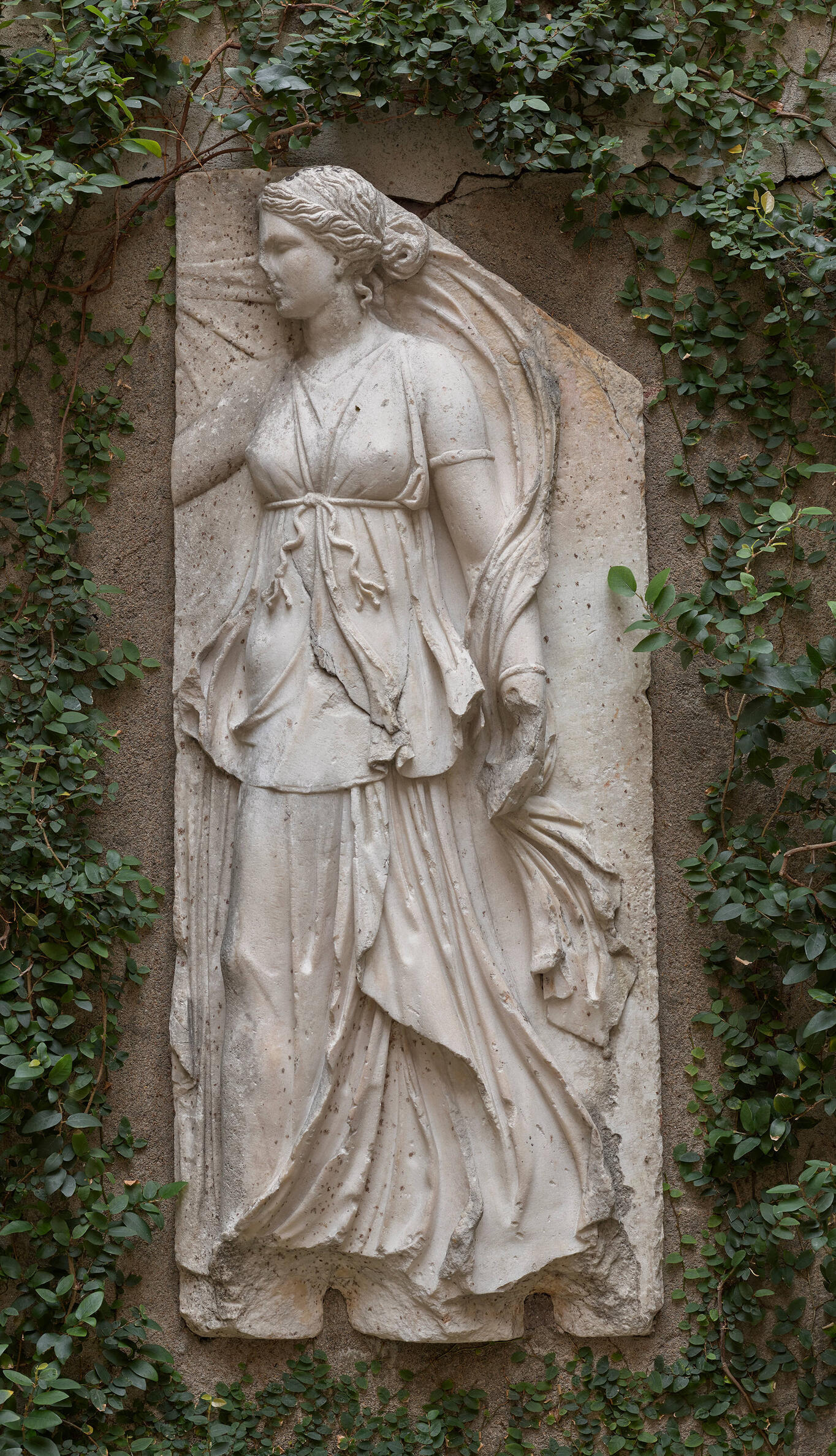 A high relief marble sculpture of a maenad facing left, with wavy hair pulled up on the back of her head and wearing flowing, draped fabric, which is secured with a tied cord below her breasts.