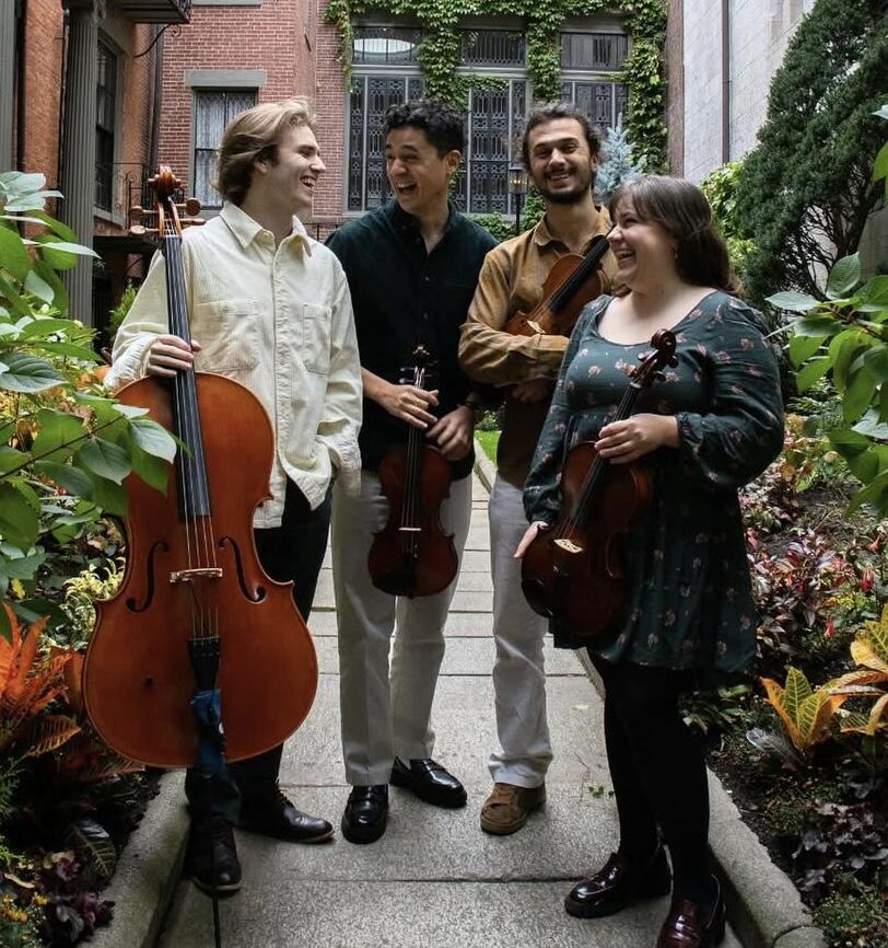 Four young adults standing outdoors holding string instruments smililng. 