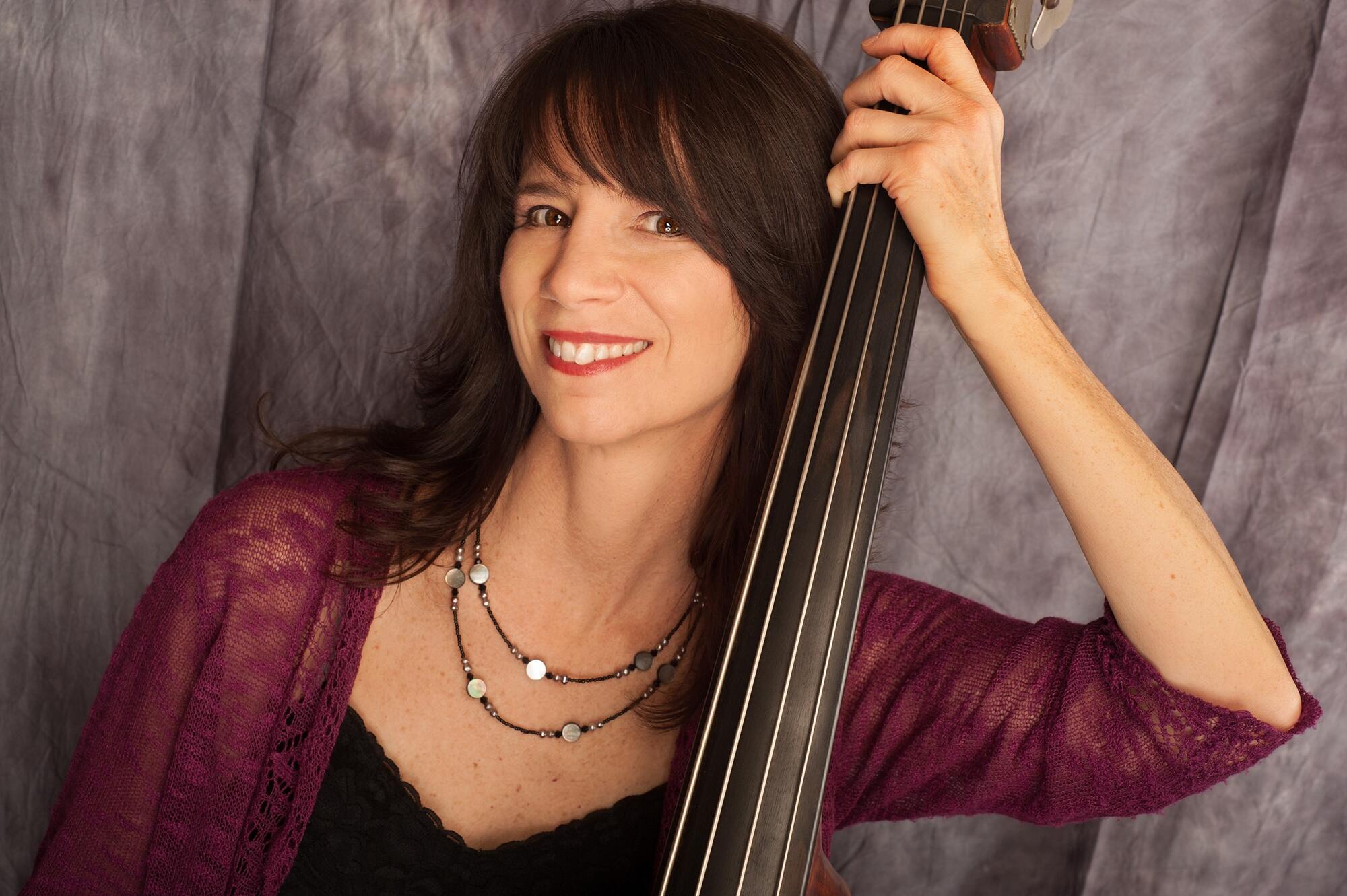Young woman with long brown hair, smiling leaning against a string instument. 