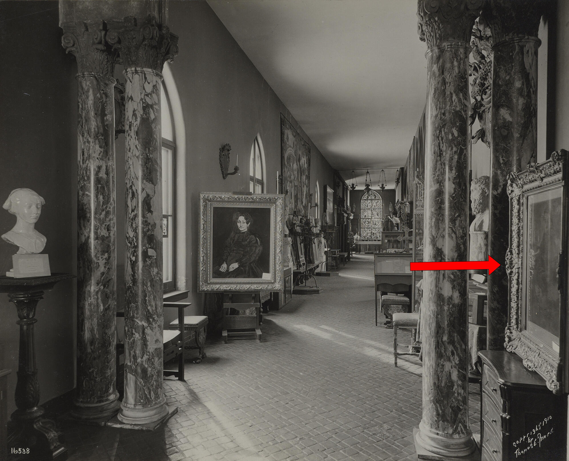 : A black and white photograph of a long narrow gallery. A red arrow points to a painting on the wall on the right side.
