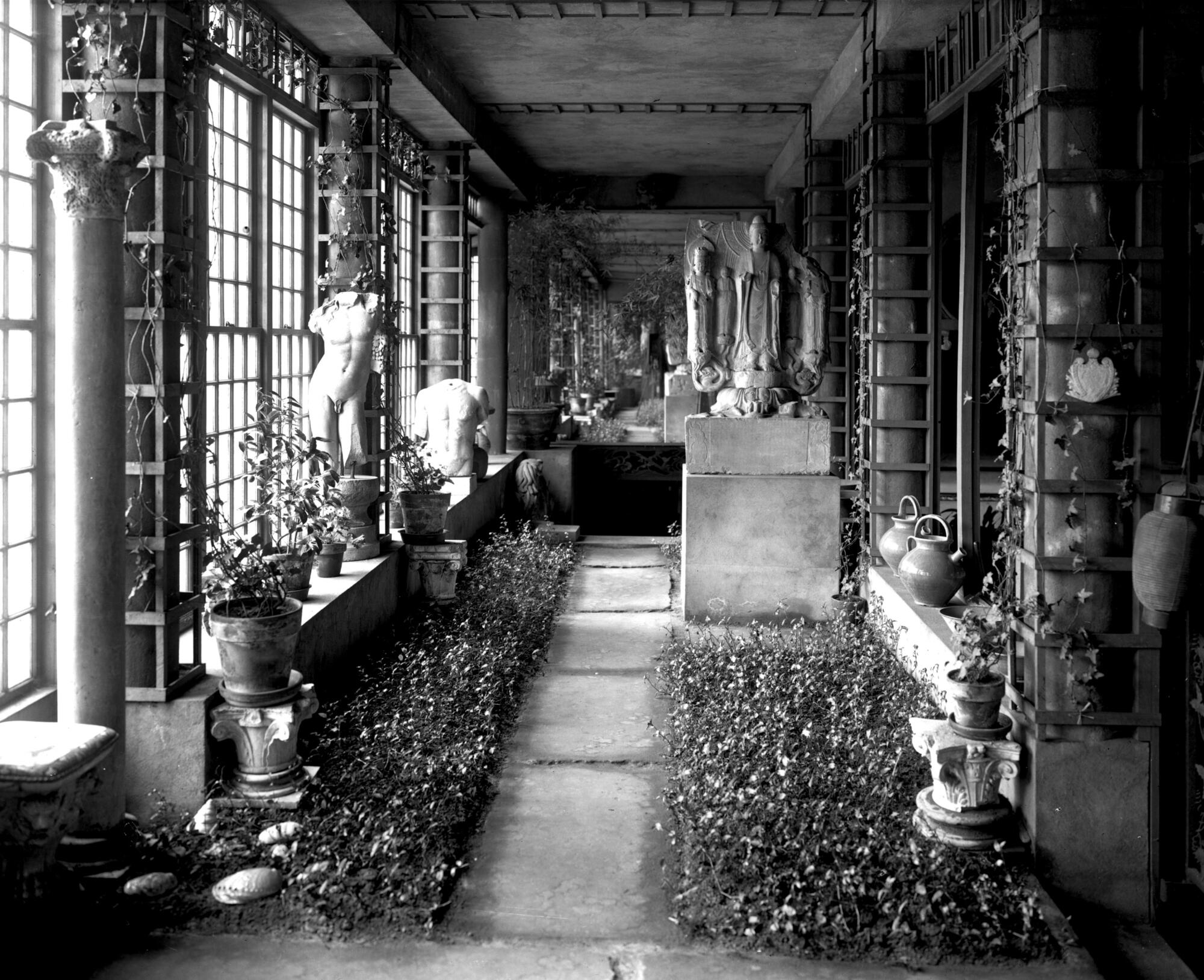  A black and white photography of the south end of the Chinese Loggia at the Gardner, with flowers lining a stone path that flows from the foreground to the background, and sculptures on either side of the path, including a limestone Buddha votive stele on the right and the torso of Dionysus on a stand against the windows to the left.