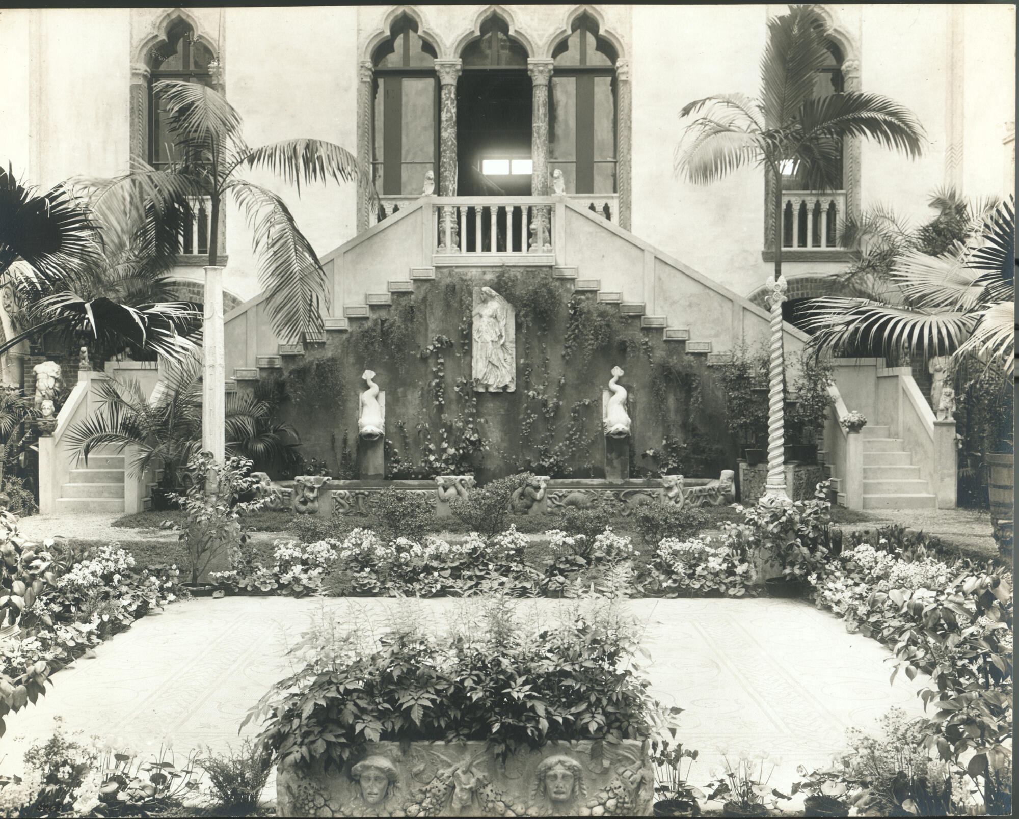 A black and white photograph of the south wall of the Courtyard at the Gardner, featuring a sarcophagus and mosaic and surrounded by flowers in the foreground and a fountain in the background framed by two sets of stairs leading up to a balcony with arched windows.