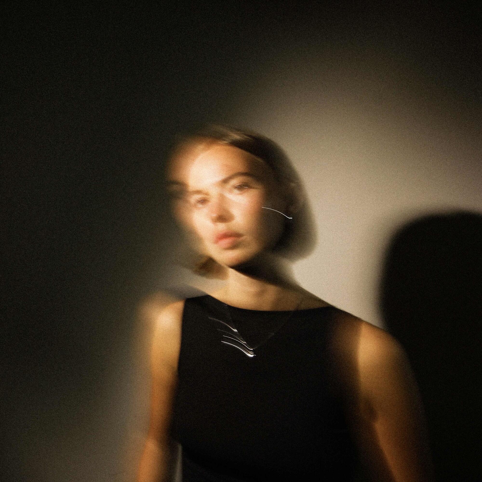 Blurred photo of a young woman in a black dress, short hair looking directly at the camera. 