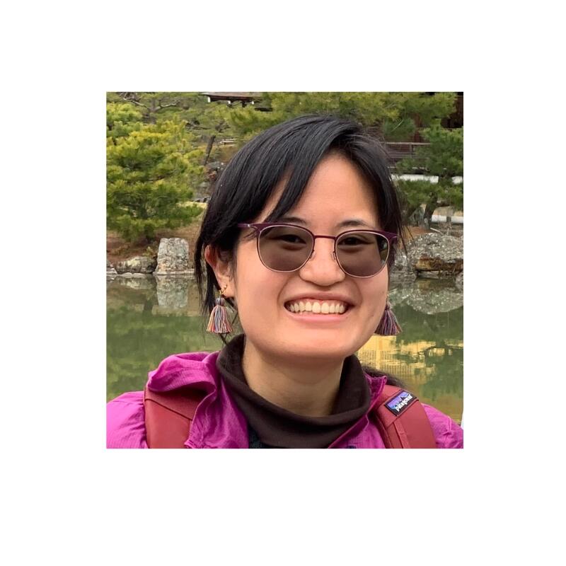 Young Asian woman with short dark hair, sunglasses standing in front of a pond and trees, smiling at the camera. 