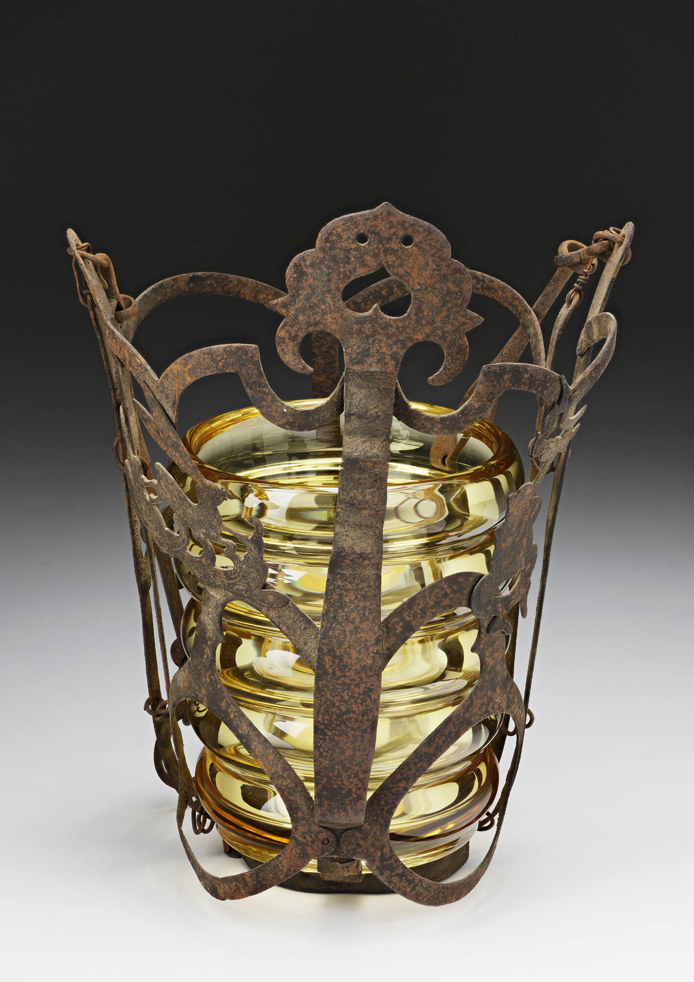 An iron cylindrical basket holding a yellow glass vessel.