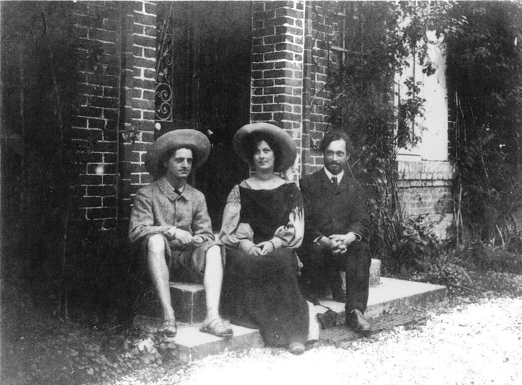 A black and white photograph of siblings Raymond Duncan, Isadora Duncan, and Augustin Duncan. Raymond Duncan wears a wide brimmed hat, suit jacket, and pants pushed up to his knees. He wears sandals on his feet.