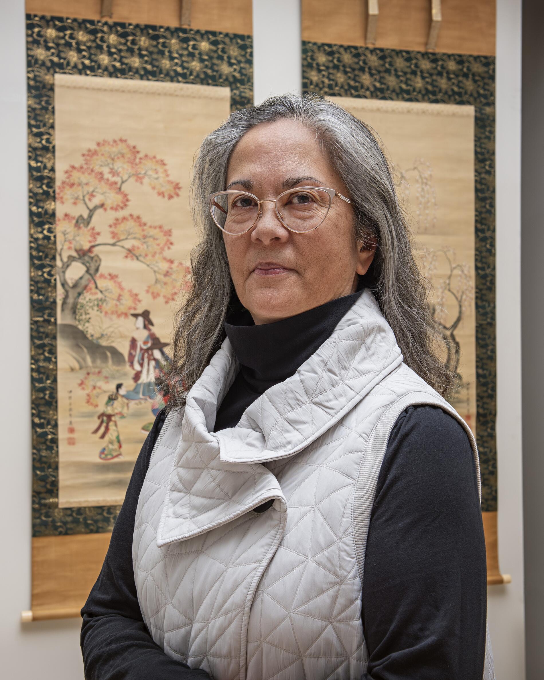 Asian woman with long grey hair and glases, looking directly at the camera, standing in front o 