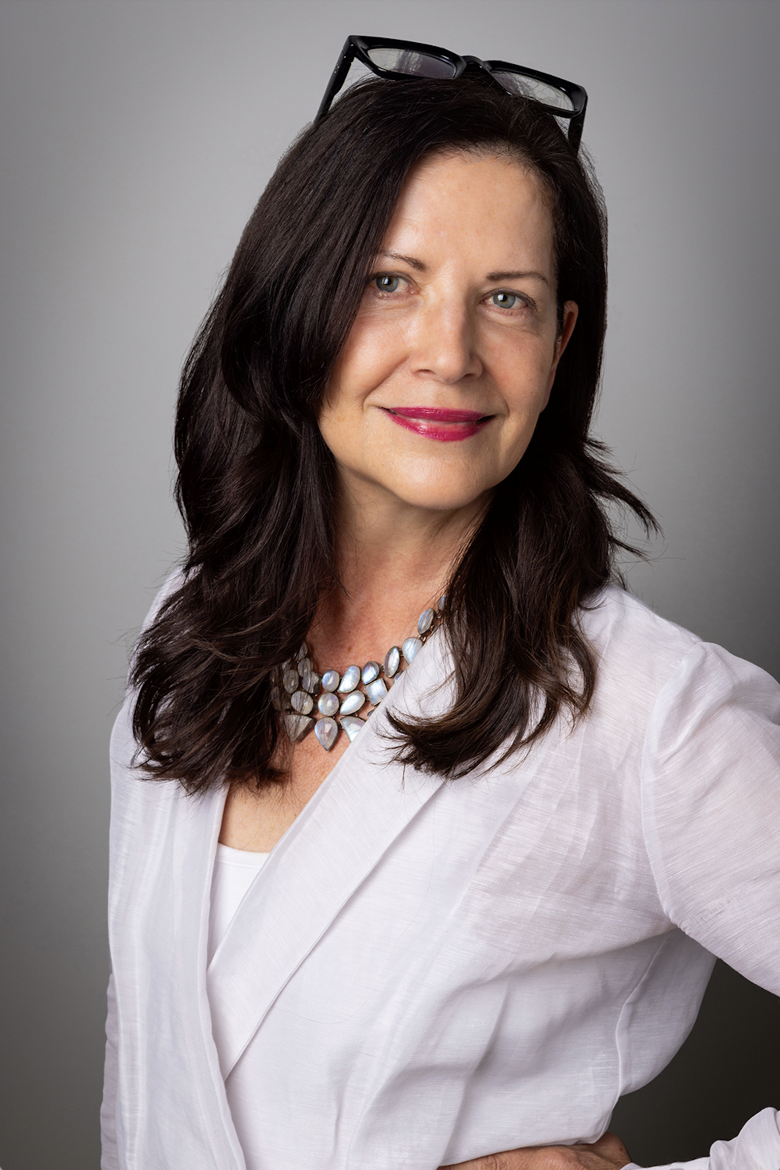 White woman with long brown hair with glasses on top of her head, weaing a white top and large beaded necklace