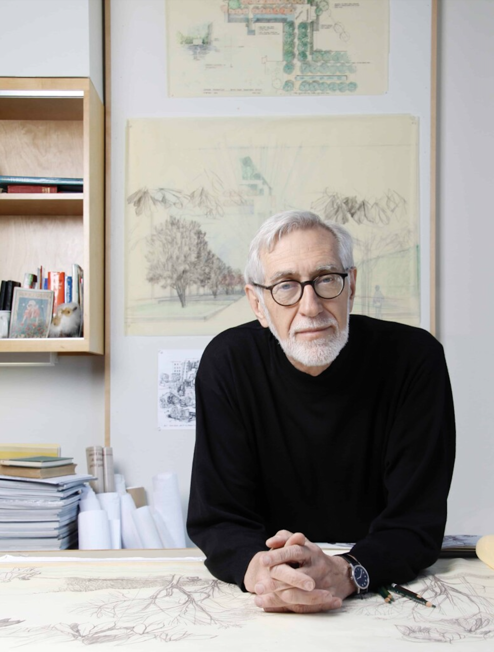 White male with short, silver hair and beard wearing a black sweater leaning on a desk with landscape designs on the wall behind him.  