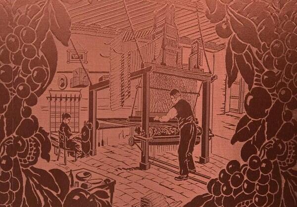 A red textile with a woven pattern depicting a family silk manufacturer; a man operates a 19th-century jacquard loom in the foreground while a woman spins silk yarns in the background. 