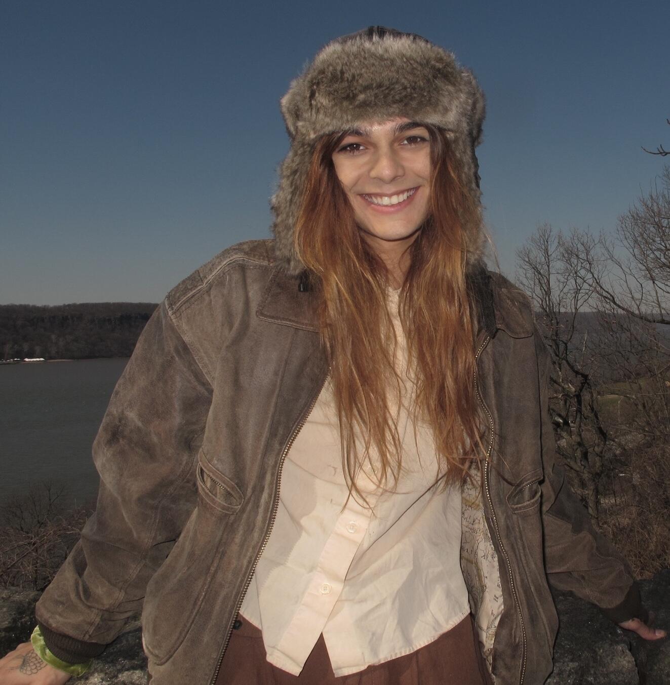 A white person with long, light brown hair with a fur had and jacket on smiling at the camera
