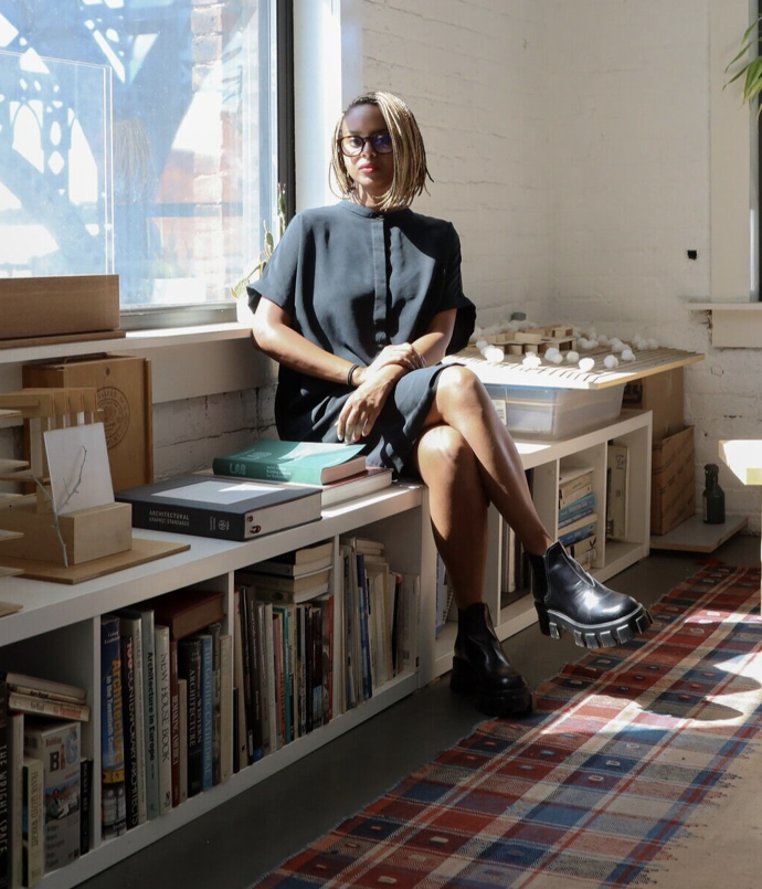 African American woman with blonde shoulder length braids and glasses, wearing a black dress and is sitting on a short bookcase. 