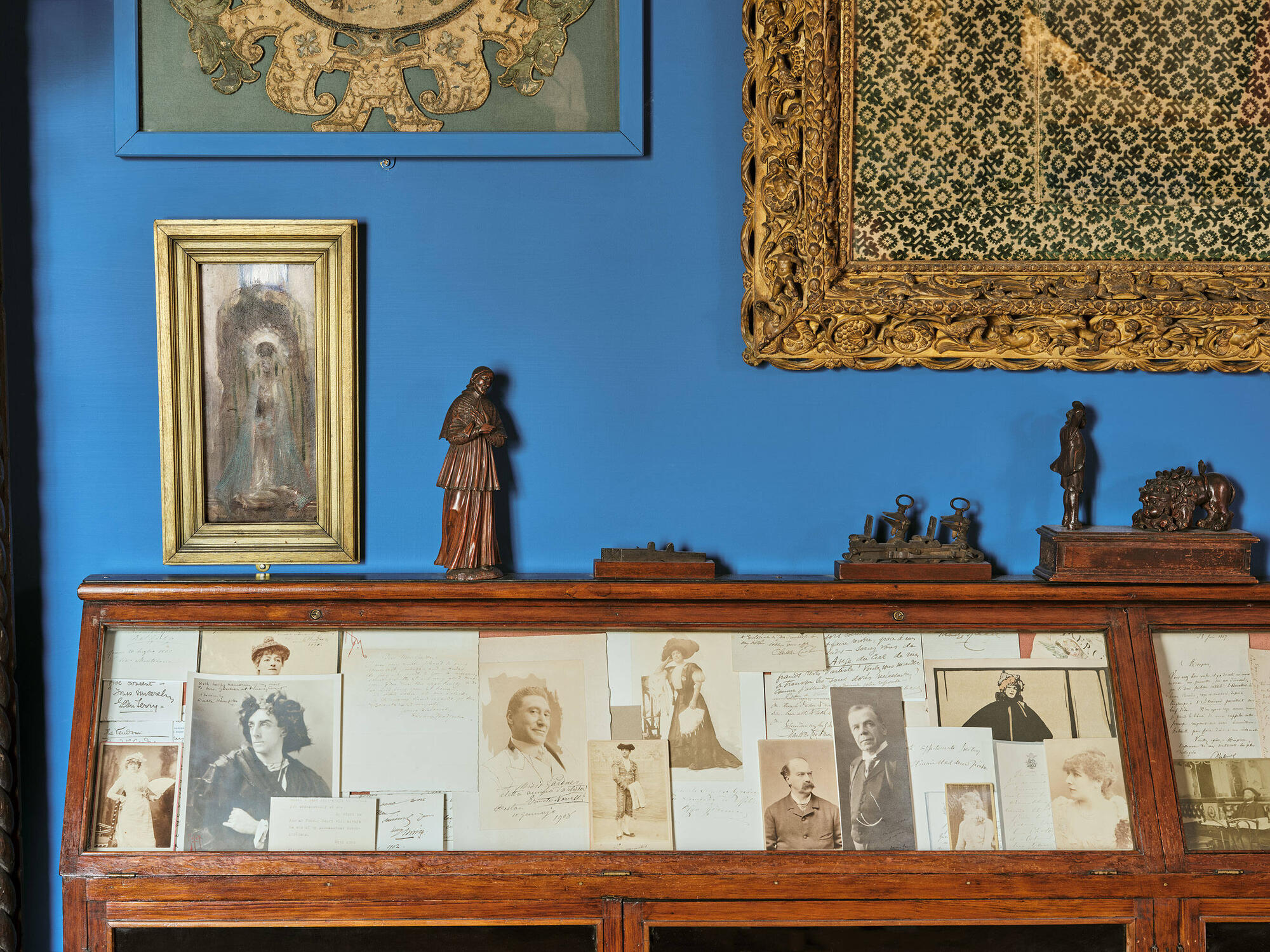  Detail of a wood and glass display case filled with overlapping photographs, memorabilia and letters. The woodcut print of Sarah Bernhardt is partially covered up by photographs and documents. The display case sits in front of a bright blue wall with small sculptures displayed on top of the case and paintings hung above. 