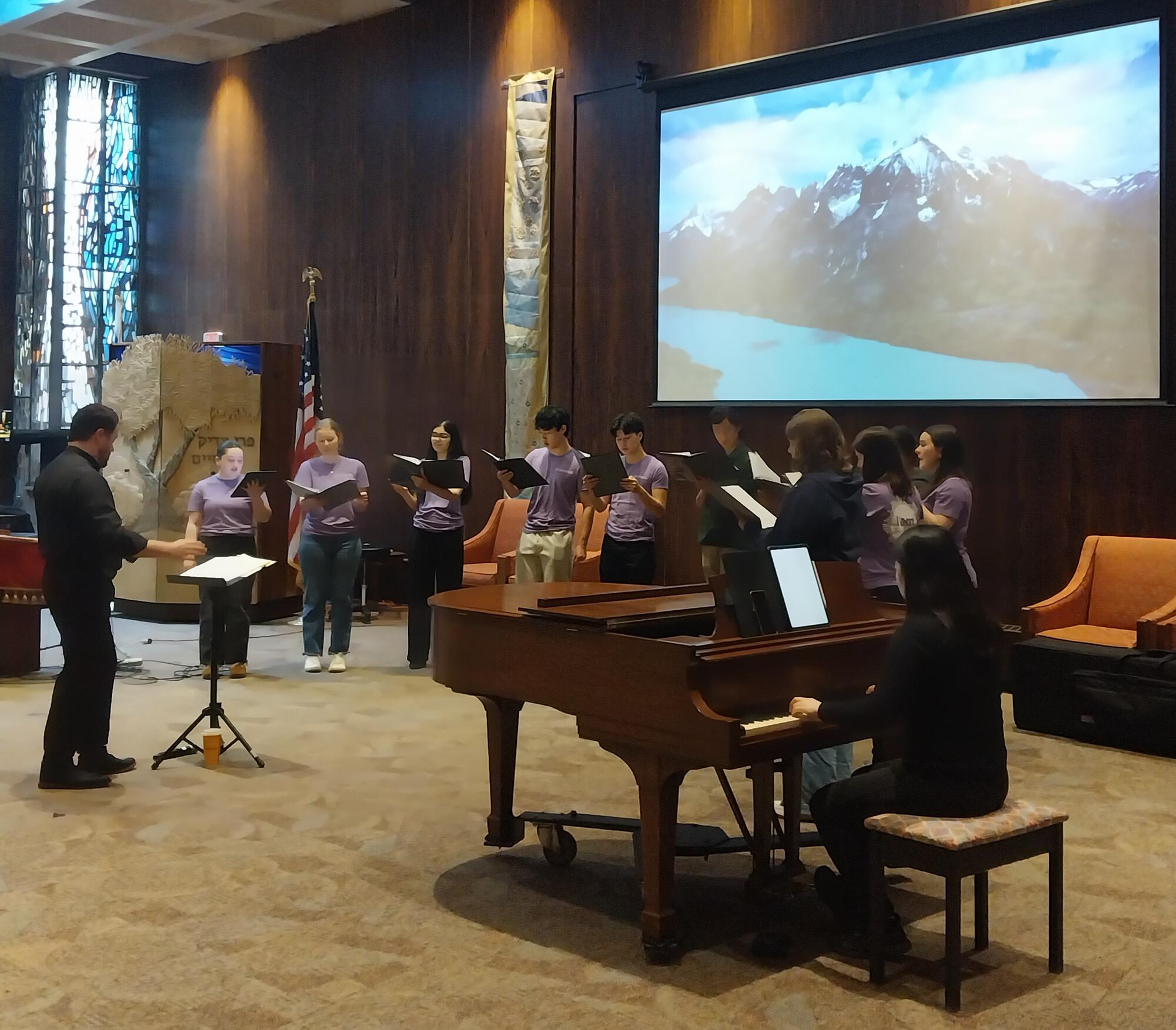A group of youth standing in front of a large screen with song books in their hands with an adult conducting them and another adult playing the piano