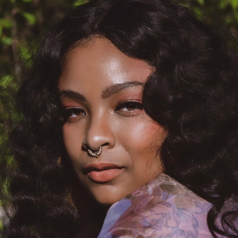 Photo of an African American with long dark wavy hair, a nose ring, looking straight at the camera.