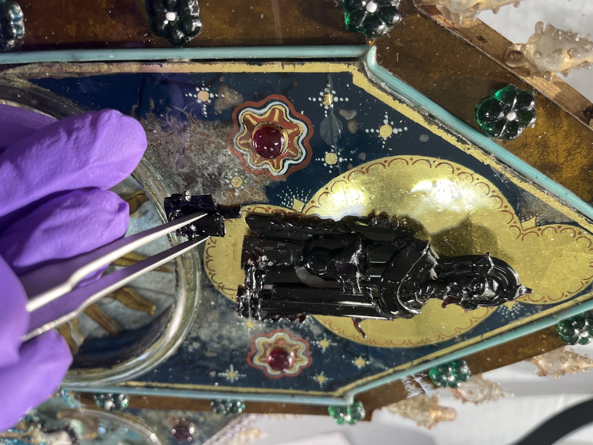  A conservator’s gloved hand holds a pair of tweezers with a small piece of dark purple glass. The conservator is carefully disassembling a glass figure of God on the sculpture as part of the conservation treatment.