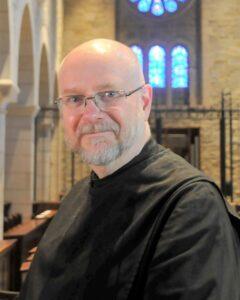 White, bald man with a grey goatee and glasses wearing a black cassock