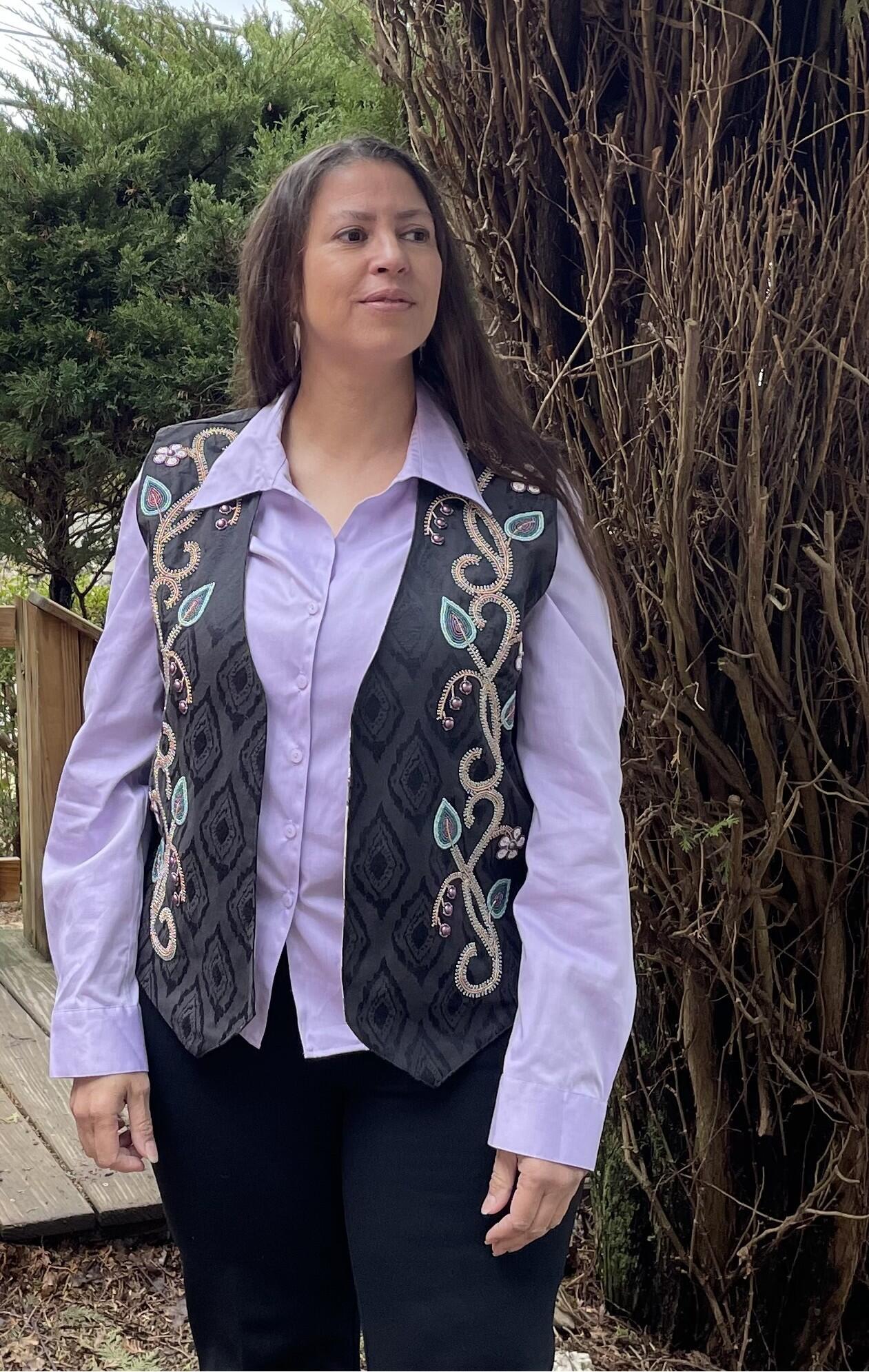 A woman with long dark hair wearing an embroidered vest with a purple blouse and dark pants standing in front of large dried plants outdoors. 