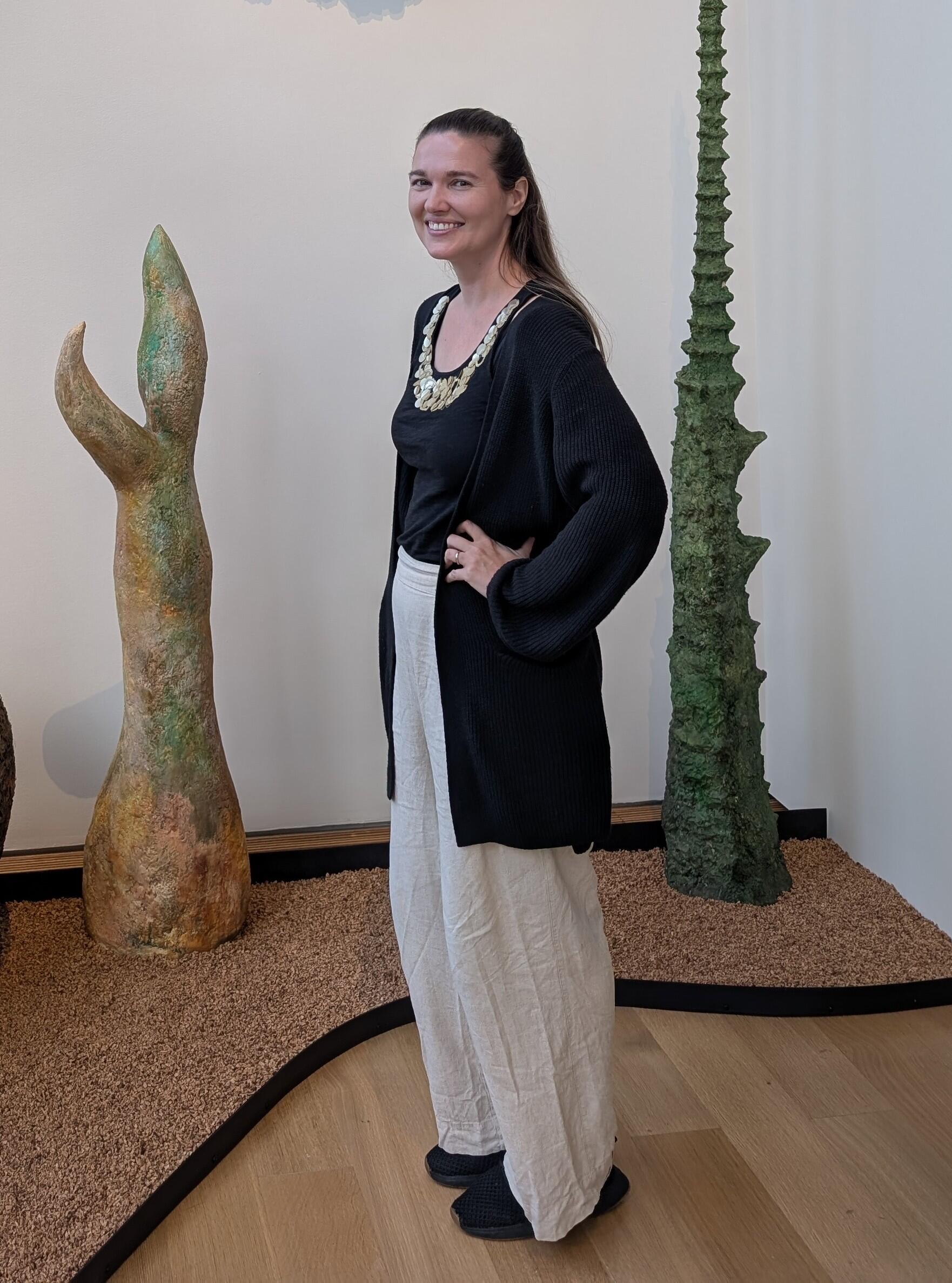 A white woman with long, straight dark hair wearing white slacks, a black shirt and long black, opened sweater standing in front of art pieces