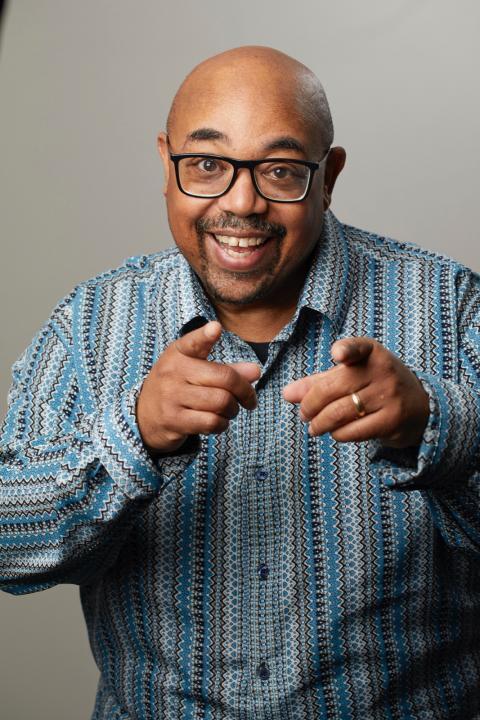Bald African American man with glasses and a big smile pointing at the camera 