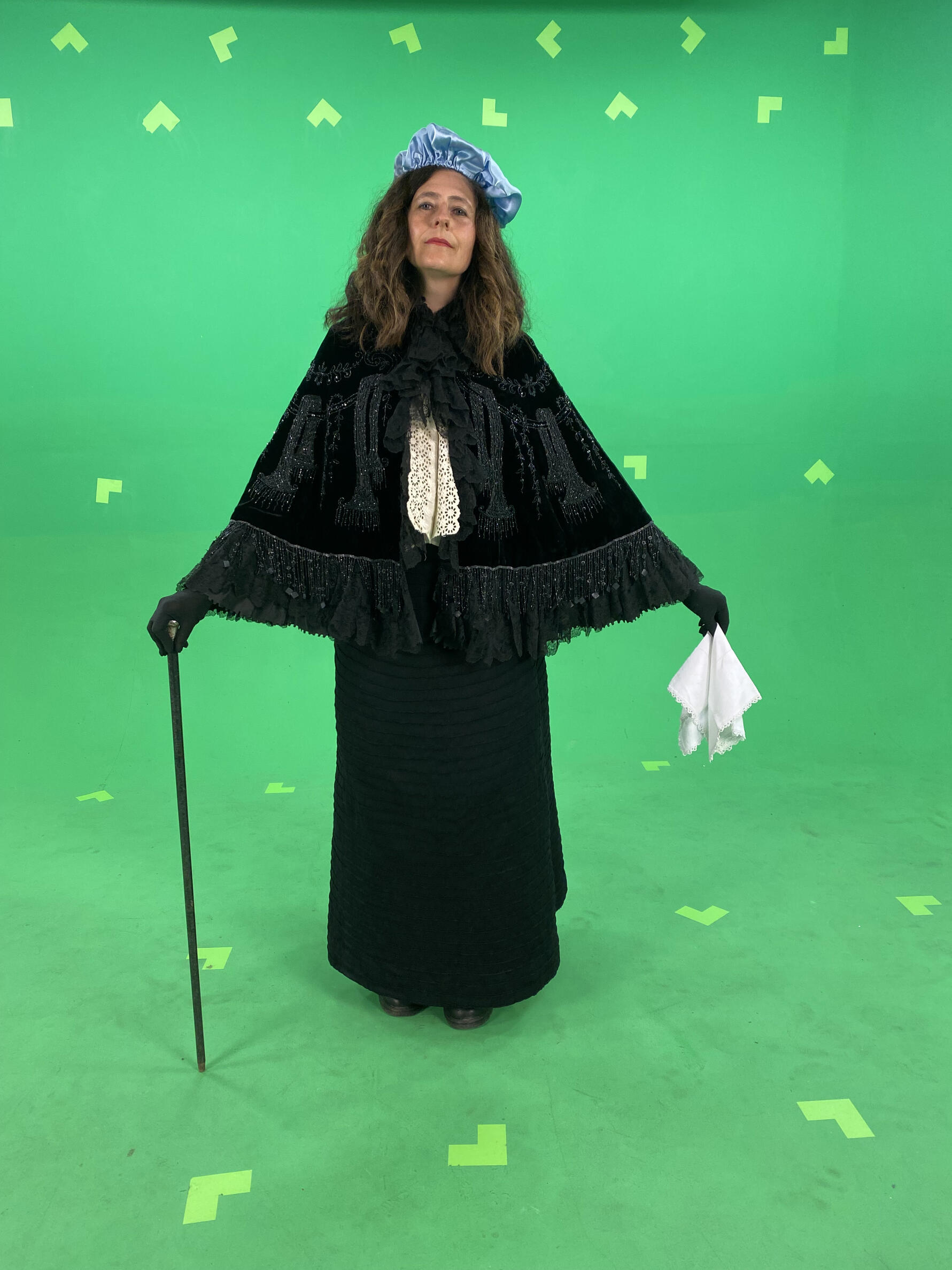 The author, a middle-aged white woman with shoulder-length brown hair, wears the historic 1890s outfit that is similar to what Sarah Bernhardt wears in the woodcut print—black cape, white blouse, long black skirt, light blue hat and black gloves. She strikes a similar pose with a white handkerchief in one hand and thin black cane in the other. She stands in front of a green screen to shoot a video clip.