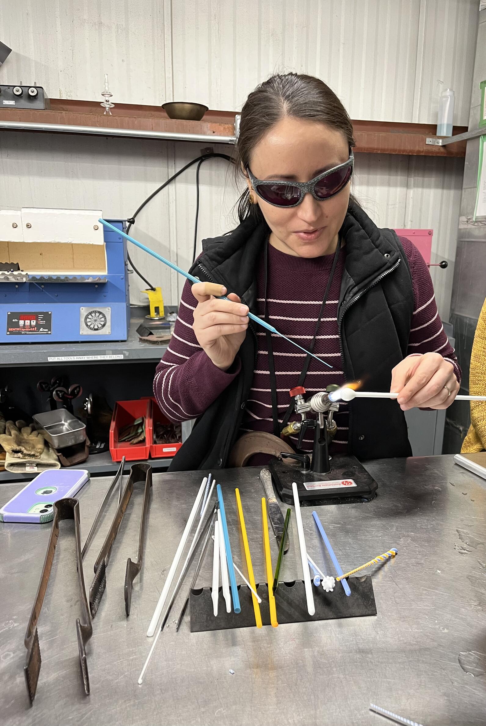 An artist wearing dark glasses to protect her eyes holds a thin rod of glass in each hand and works them over a flame torch that is mounted to the workbench. Additional glass rods and tools lay on the workbench nearby.