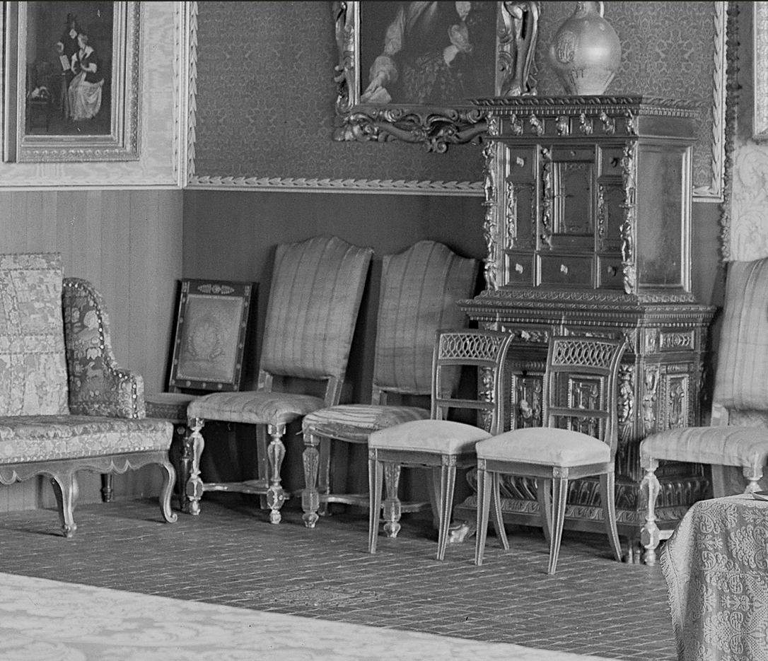 A room with European old-master paintings and furniture arranged eclectically. The lower half of the image shows a line of mismatched chairs. 