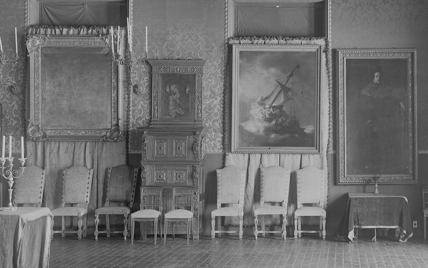A room with European old-master paintings, sculpture, and furniture arranged eclectically. The lower half of the image shows a line of chairs, one with striped ombre embroidery, the rest covered in floral damask. 