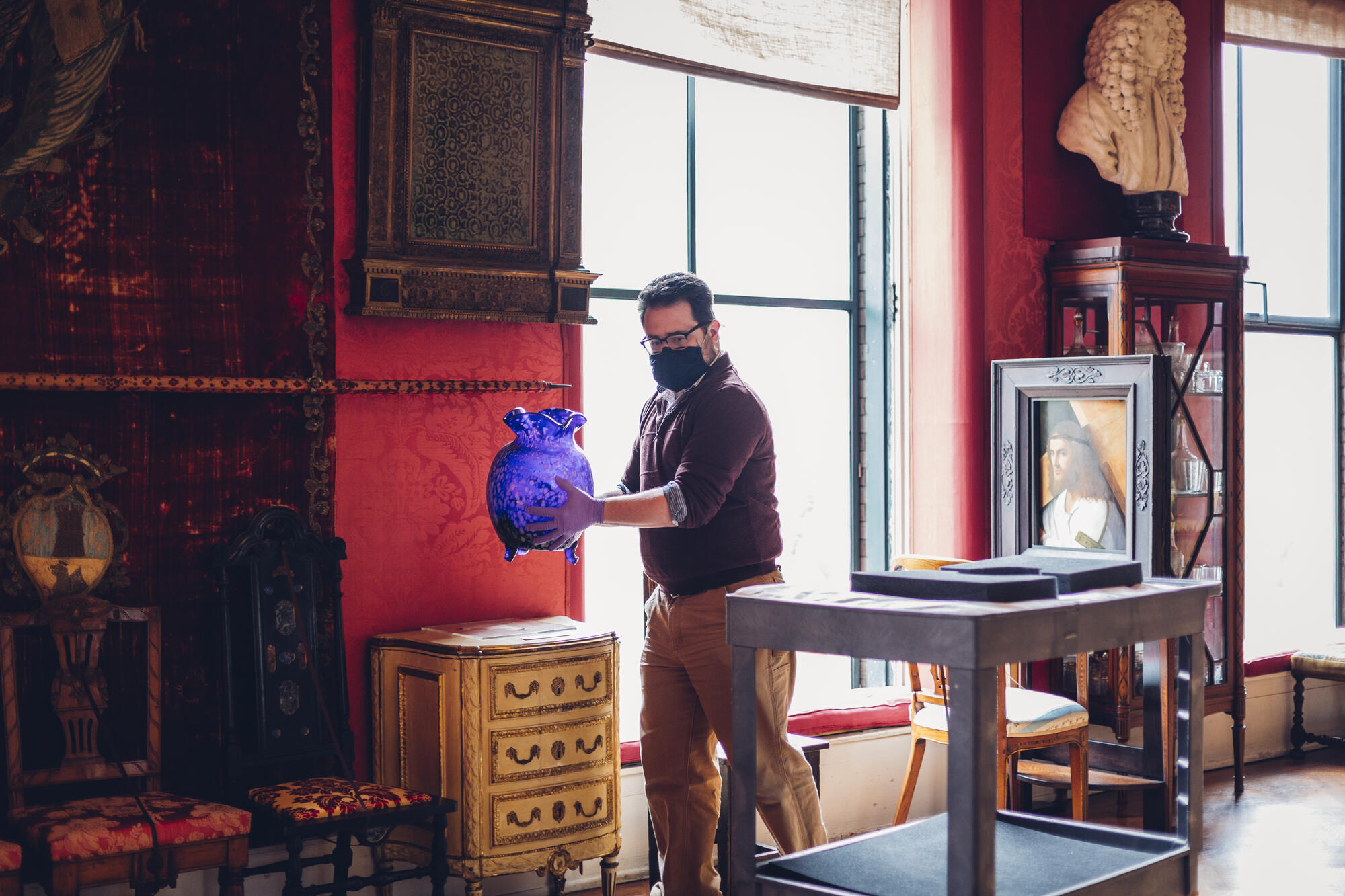 An art handler carefully moves a large blue and white speckled glass vase from a transport cart, back onto display in the Titian Room gallery.