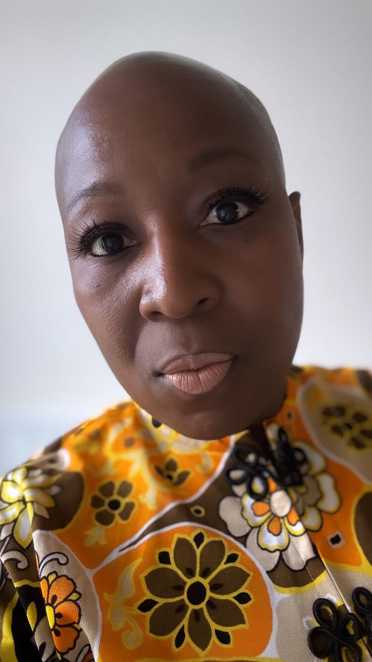 A close-up of a person with a shaved head, wearing a vibrant, patterned shirt with orange and brown floral designs. The person's facial expression appears neutral, with a slight hint of a smile. The background is plain and light-color. The shirt has a collar and features intricate floral patterns that stand out against the light background.