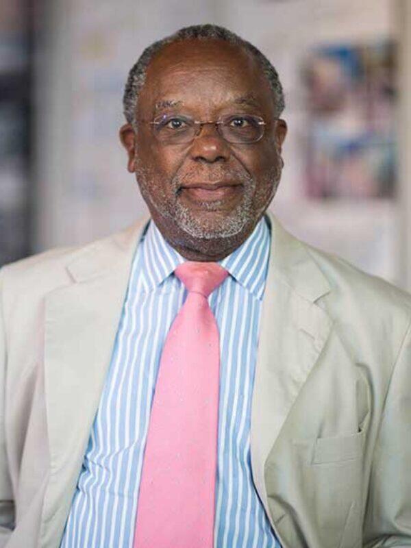 African American man with short, grey hair and goatee, glasses wearing a blue and white striped shirt, a pink ties and a tan suit jacket