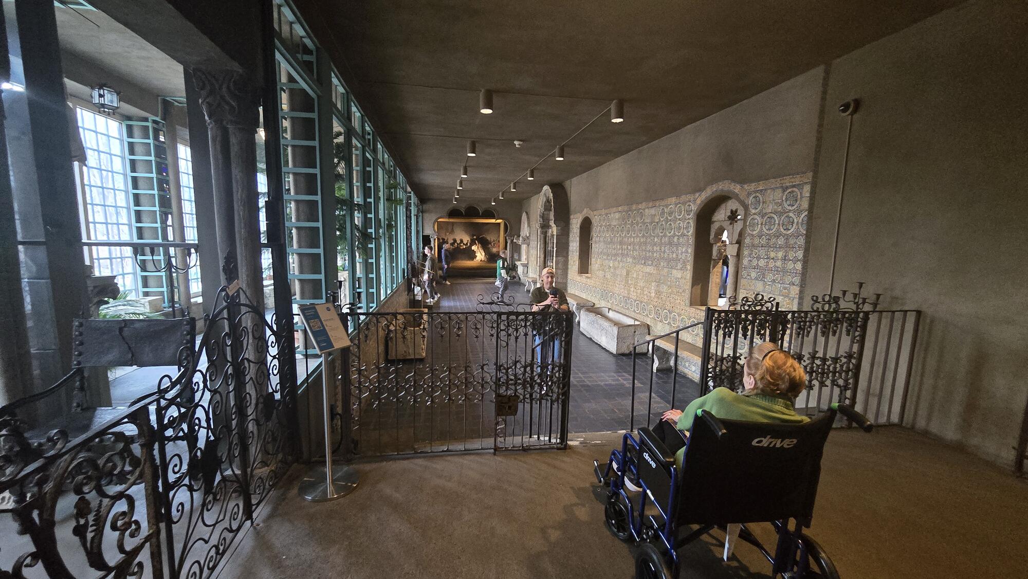 A visitor who uses a wheelchair poses at the top of the stairs in the Spanish Cloister gallery while having their photo taken. At the end of the gallery a large painting of a flamenco dancer, El Jaleo, by John Singer Sargent, is displayed.