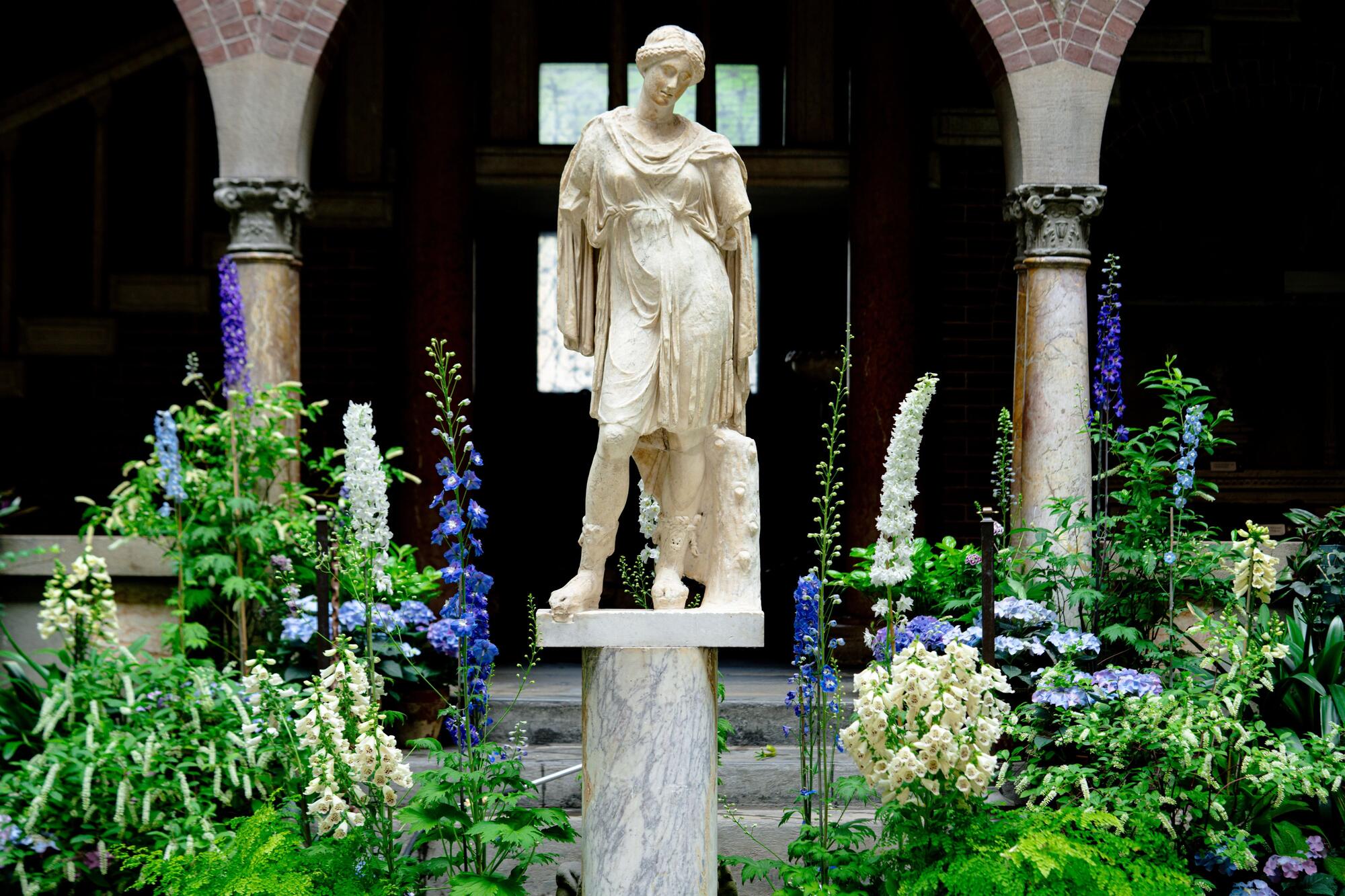 A statue of a woman on a pedestal surrounded by talk stems of white and blue flowers in the Courtyard. 