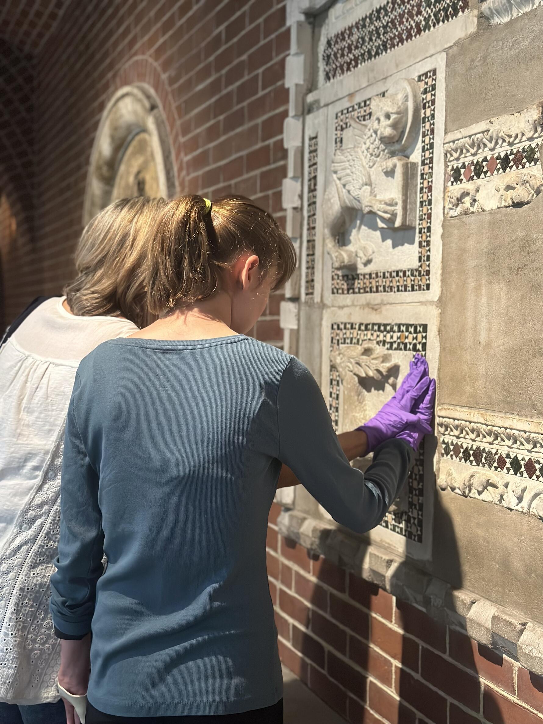 A museum teacher guides a visitor with blindness in touching a marble sculpture set into the brick wall of the museum. They both wear purple nitrile gloves while touching the sculpture.
