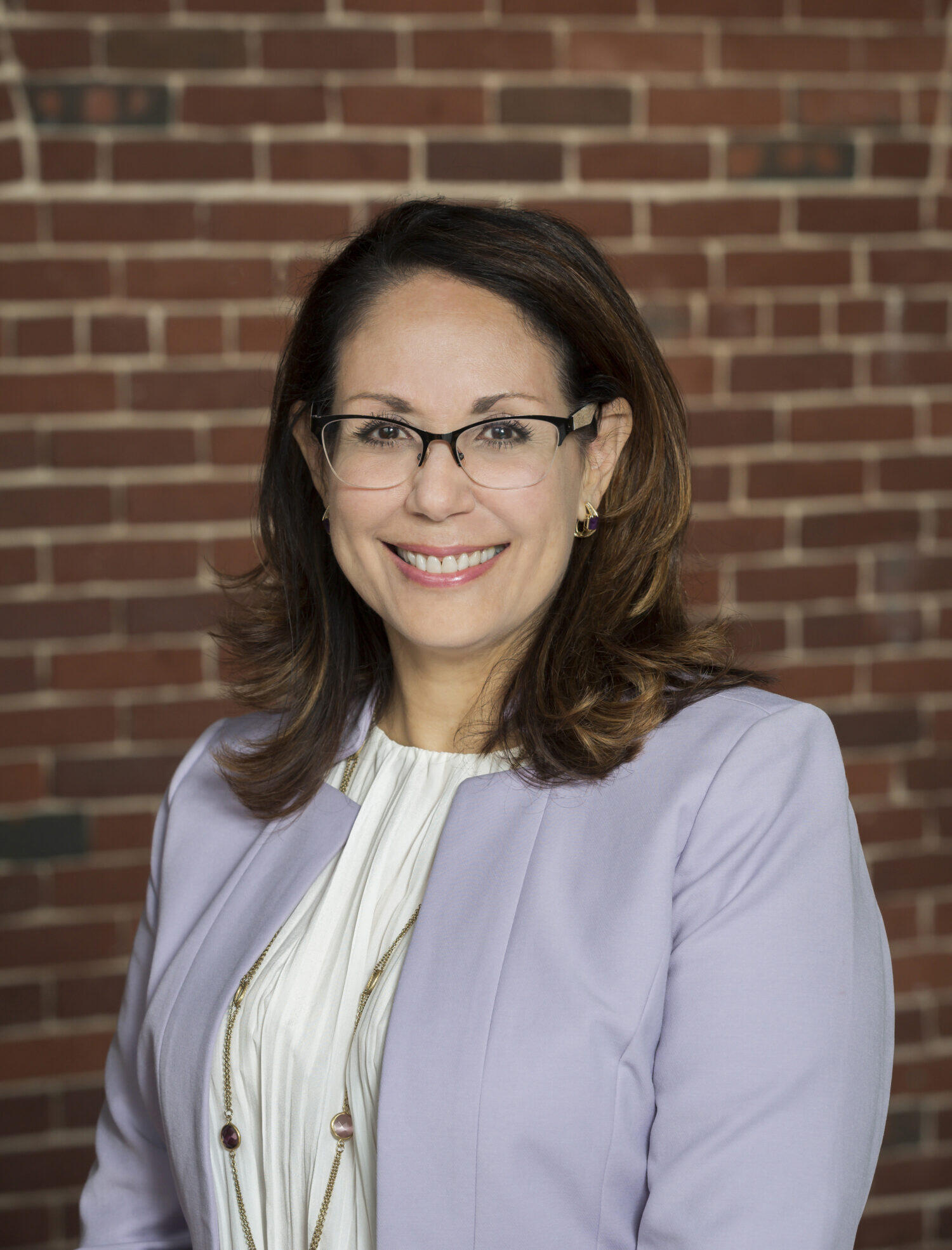 Caucasian woman with shoulder length brown hair with glasses wearing white blouse and lavender jacket smiling at the camera. 