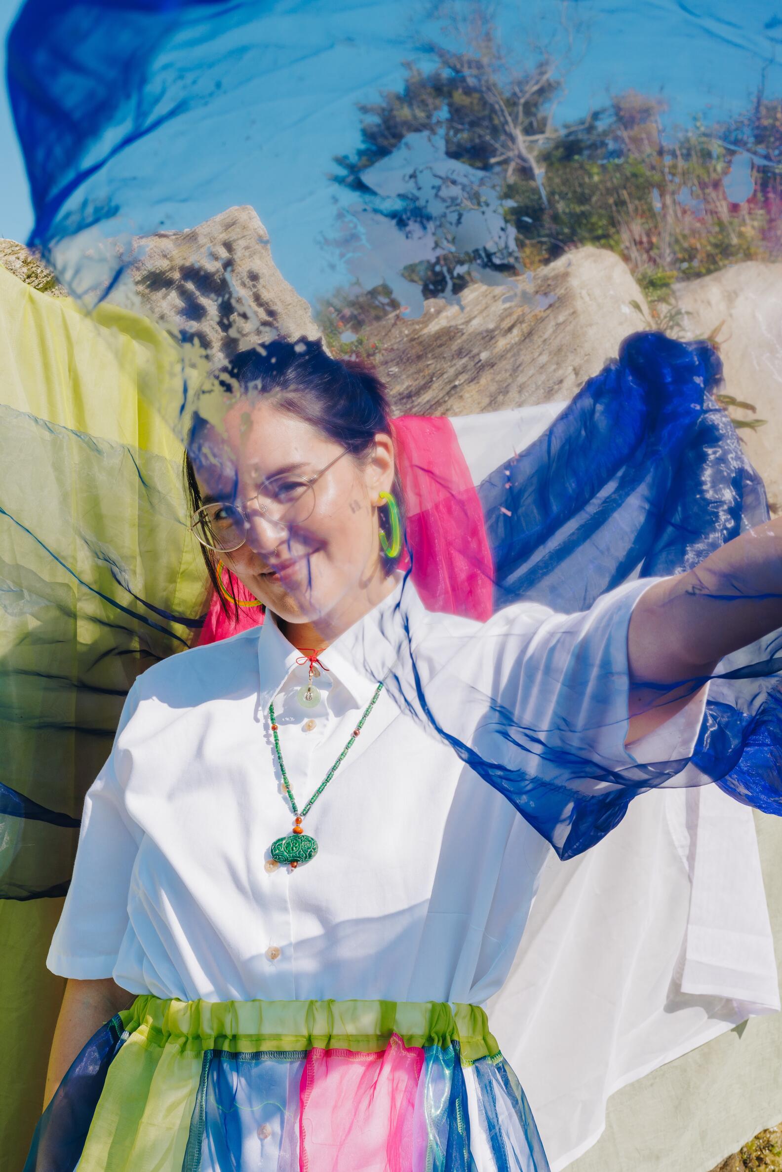 Young Asian woman with her hair pulled back wearing glasses, a shirt and colorful skirt waving a light weight blue scarf around her. 