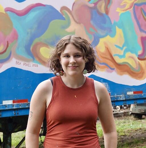 Young woman with chin-length reddish, curly hair weaing an reddish tank top standing in front of multicolored mural smiling at the camera